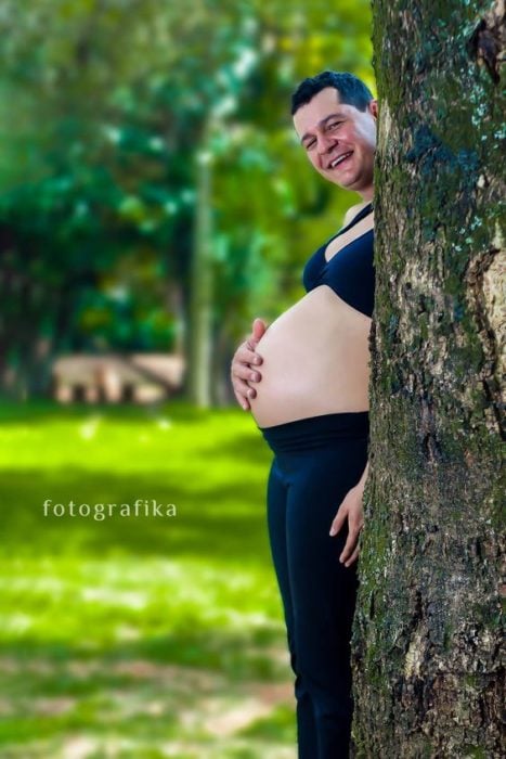 mujer embarazada se oculta tras un árbol, solo se ve su panza y sale la cabeza de su esposo
