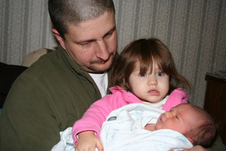 padre carga a su hija que a su vez carga a la bebé recién nacida