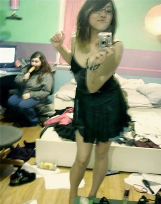 muchacha se toma una selfie en el espejo, mientras atrás su amiga come una salchicha y la habitación está desordenada